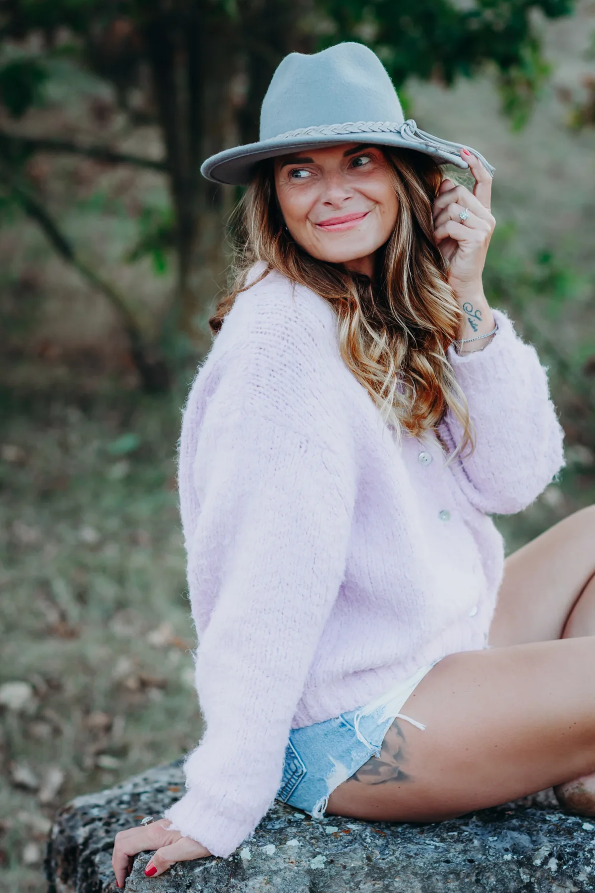Alexandra Kohler mit Hut sitzt lächelnd auf einem Stein in der Natur, in fliederfarbener Strickjacke